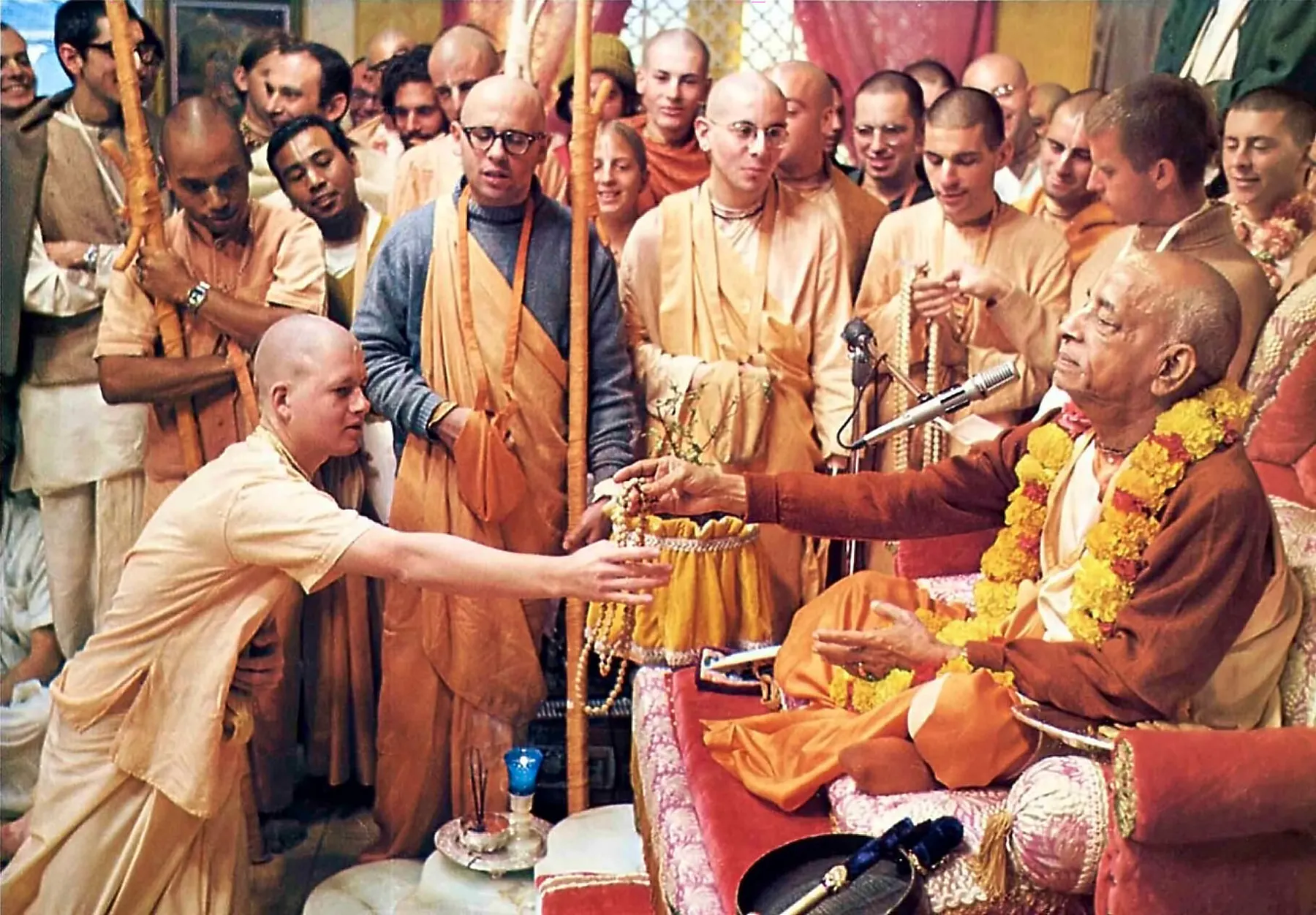 Srila Prabhupada offering initiation beads to a disciple, surrounded by devotees